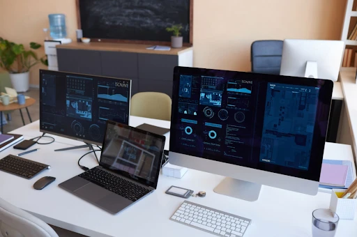 A bright, modern technology support office workspace with desktop monitors and a laptop displaying live network analytics, systems infrastructure monitoring, and data dashboards.